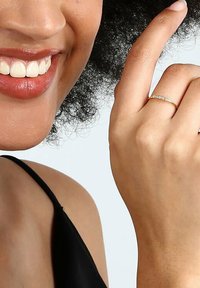 Guld ring med små klara stenar, som bärs på en hand med neutral hudton, bredvid ett leende ansikte och lockigt svart hår, mot en ljus bakgrund.