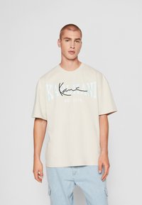 T-shirt en coton beige à manches courtes avec "Kahlani" imprimé en bleu et noir, col rond, porté avec un pantalon cargo bleu clair.