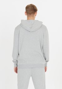 Man som bär en ljusgrå huvtröja och matchande sweatpants, står vänd bort mot en enkel vit bakgrund.