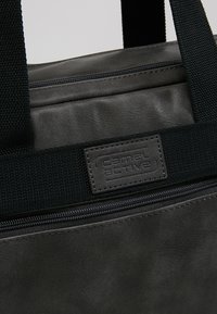 Schwarze Stofftasche mit glatter Oberfläche, ausgestattet mit einem Reißverschluss oben und einem strukturierten schwarzen Trageriemen. Enthält ein rechteckiges Logopatch.