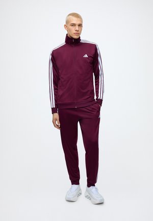Joven de pie con un chándal Adidas marrón oscuro con rayas blancas en las mangas y los pantalones, combinado con zapatillas deportivas blancas.