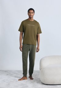 Λαδί βαμβακερό T-shirt με κοντά μανίκια και κείμενο λογότυπου, συνδυασμένο με αντίστοιχο παντελόνι φόρμας, που βρίσκεται σε ανοιχτό γκρι χαλί.