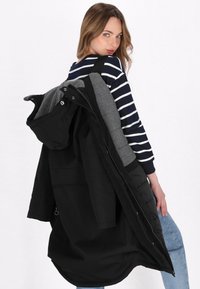 Cappotto a strati nero e grigio con cappuccio, dotato di zip e tasche, indossato sopra un top a righe blu e bianche, che mostra una vestibilità comoda.