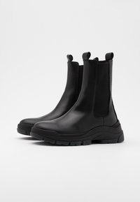 Botas de tobillo de cuero negro con paneles laterales elásticos, tiradores y suelas de goma gruesas. Textura suave con un diseño elegante y aerodinámico.