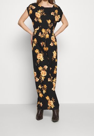 Femme portant une robe maxi noire à motif floral orange et des bottines marron, debout les mains sur les hanches devant un fond blanc.