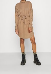 Robe à manches longues, couleur sable, avec une taille ceinturée et un ourlet arrondi, portée avec des bottines noires à lacets et des chaussettes rehaussées d'or. Texture lisse.