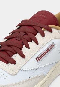 Gros plan sur une basket Reebok blanche et beige avec des lacets rouges et un logo, montrant du cuir texturé et des détails cousus.