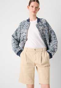 Veste bomber florale dans des nuances de bleu, avec une fermeture éclair et des poignets côtelés, associée à un short beige plissé.