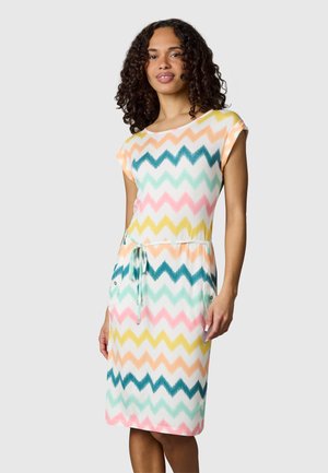 Kobieta z kręconymi włosami, ubrana w kolorową sukienkę o długości do kolan z krótkimi rękawami, z wiązaniem w talii i wzorem zygzaków.