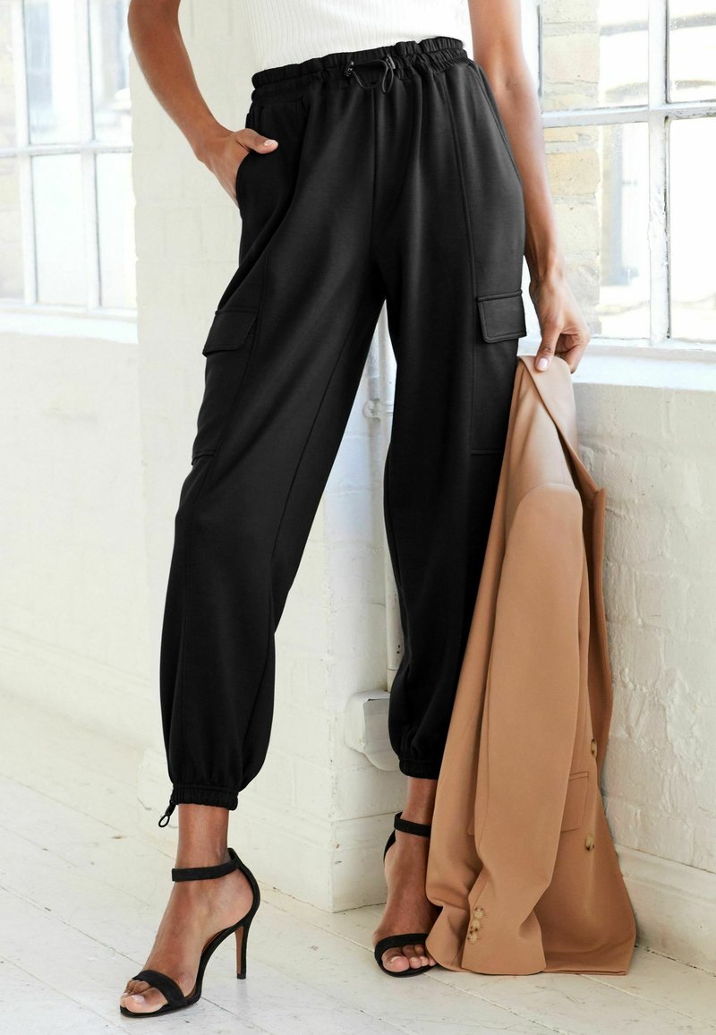 Femme portant un pantalon jogger cargo noir et des sandales à talons noires, tenant un blazer beige près d'une fenêtre dans une pièce lumineuse.