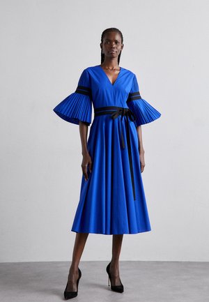 Blå plisseret kjole med en dyb V-hals, sorte stribede detaljer og et sort bælte. Ærmerne er brede og flagrende, hvilket skaber en svævende silhuet.