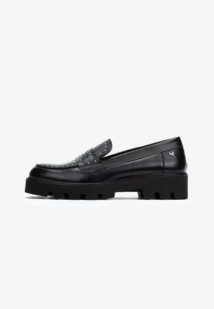 Mocassin en cuir noir avec semelle épaisse à crampons, clous métalliques sur le dessus et la bande, et un petit emblème argenté en forme d'aile au talon.