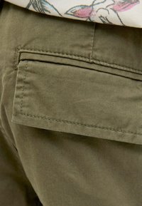 Gros plan sur une poche cargo en tissu vert olive avec rabat et coutures sur un pantalon porté avec une chemise à motif floral.