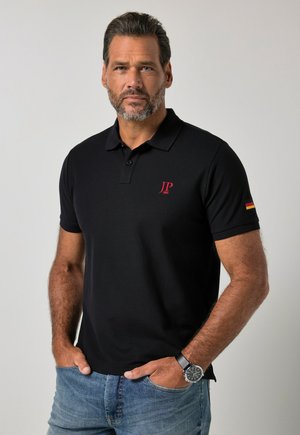 Mand med skæg iført sort poloshirt med rødt "JP 1883" logo og det tyske flag på ærmet, blå jeans og sort armbåndsur, stående.