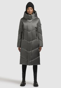 Lång, grå puffjacka med hög krage, chevrondesign och dragkedjefickor. Buren med en svart stickad mössa och svarta ankelstövlar.