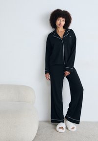 Musta pyjamapuku, jossa on pitkähihainen paita ja leveälahkeiset housut, joissa on valkoista piparkoristetta. Malli pitää yllä pehmeitä valkoisia tossuja ja seisoo vaalealla matolla.