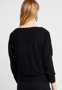 Zwarte, gebreide trui met een losse pasvorm, wijde halslijn en driekwartmouwen. De textuur lijkt zacht en lichtgeribbeld.