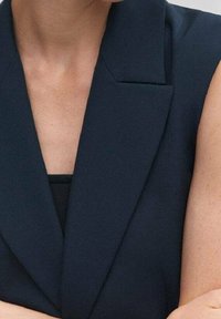 Námořnický modrý bezrukávový blazer s ostrými klopami a přiléhavým střihem. Hladká textura, minimalistický design a hluboký výstřih do V.
