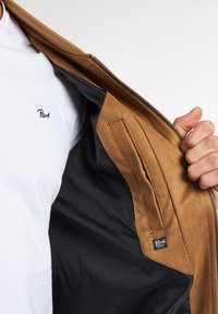 Giacca in suede marrone con foderatura nera liscia, dotata di chiusura con zip e tasca interna. È visibile una camicia bianca con ricami neri.