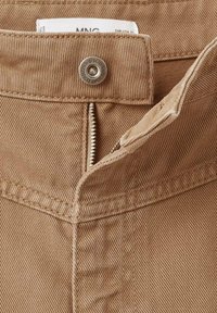 Beige katoenen broek met een zichtbare ritssluiting, metalen knoopsluiting en label. De stof heeft een subtiele ribbelstructuur.