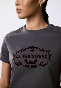 Graues Baumwoll-T-Shirt mit einem burgunderfarbenen Druckdesign, das "LA PARISIENNE EST. 1982" und verzierte Details zeigt. Mit umgekrempelten Ärmeln und Rundhalsausschnitt.