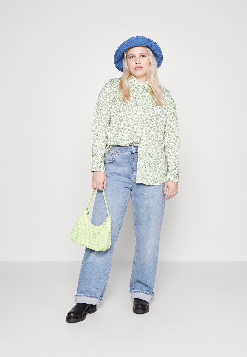 Camicia verde a fantasia floreale, jeans a vita alta blu chiaro, borsa intrecciata verde, cappello blu e stivaletti neri robusti. Sfondo semplice.