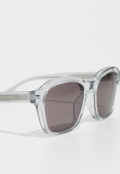 Ochelari de soare din plastic transparent, cu lentile pătrate supradimensionate, lentile gri tintate și accente argintii pe temple. Textură netedă, design modern.