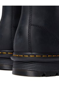 Bottes en cuir noir avec semelles épaisses à rainures et coutures jaunes distinctives le long du bord de la semelle, montrant les détails du talon et de l'arrière.