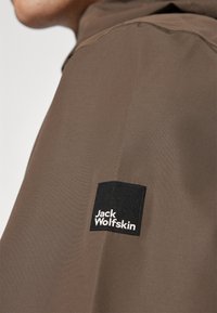 Brun jakke med en glat tekstur, med et sort kvadratisk logo-patch mærket "Jack Wolfskin" på skulderen.