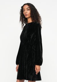 Musta samettimekko pitkillä hihoilla, vyötärö on vartalonmyötäinen ja helma levenee alaspäin. Kangas on pehmeä ja siinä on hienostunut kiilto.