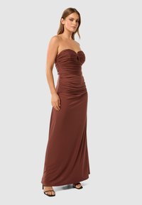 Robe maxi marron sans bretelles avec tissu froncé et détail en ouverture au niveau de la poitrine. Texture lisse et design ajusté. Sandales à talons noires.