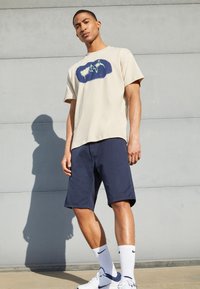 Camisola de algodão bege com um design gráfico azul-marinho, combinada com calções azul-marinho, meias brancas desportivas e ténis brancos, em pé contra uma parede cinza.