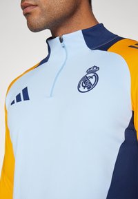 Világoskék pulóverdzseki tengerészkék és narancssárga részletekkel. Cipzáras nyakkal és a Real Madrid logójával a mellkasán. Sima textúra.