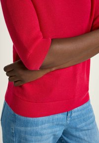 Red knitted sweater with short sleeves, textured pattern, and ribbed hem, paired with light blue jeans. Close-up view of garment and arm.