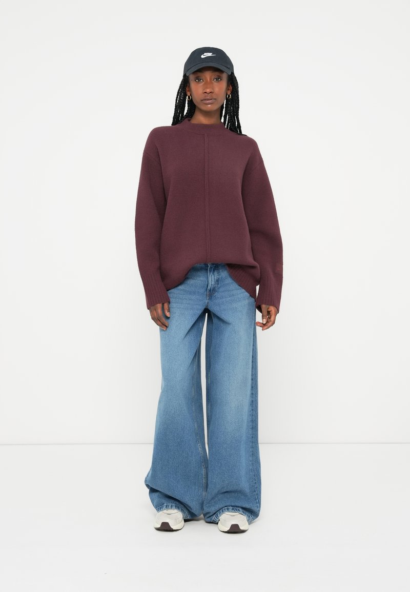 Burgundy oversized tröja med hög hals, kombinerad med ljusblå vida jeans och vita sneakers med mörka accenter, grå keps.