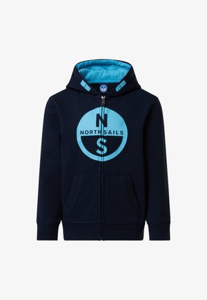 Garçon blond portant un survêtement bleu marine avec un logo bleu clair, baskets blanches, debout avec une main dans la poche, regardant vers le bas.