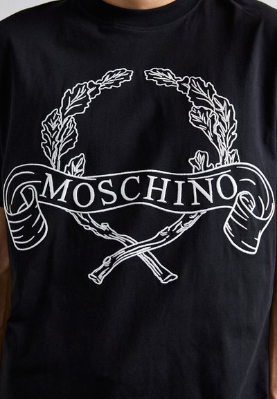 Must t-särk mustast puuvillast, millel on valge tikandiga loorberipärg ning "Moschino" tekst ribal koos ristõmbluste aktsentidega.
