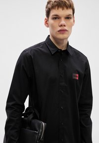 Μαύρο πουκάμισο με κουμπιά από βαμβακερό μείγμα, με δομημένο γιακά και κόκκινο-μαύρο σήμα στο στήθος. Το μοντέλο κρατάει μια μαύρη τσάντα.