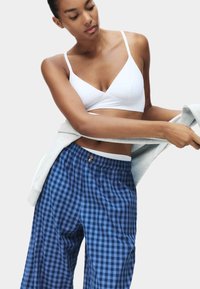 Weißer, dreieckiger Bralette und lockere, hellblaue, karierte Schlafanzughose mit elastischem Bund; Modell hält einen grauen Pullover, der über ihren Arm drapiert ist.