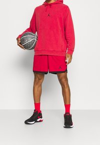Sweat à capuche rouge avec un logo noir, short rouge assorti avec des accents noirs, chaussettes rouges et chaussures de sport noires avec des détails blancs, tenant un ballon de basket.