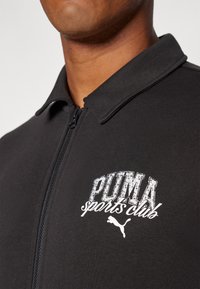 Chaqueta negra con cremallera fabricada en un tejido suave, con un cuello alto y un logo blanco que incluye el texto "PUMA sports club" en el lado izquierdo del pecho.