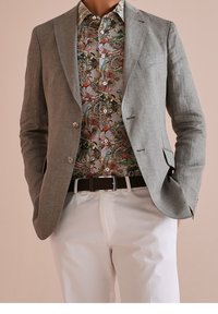 Mann trägt ein hellgraues Blazer über einem floral gemusterten Hemd mit tropischen Vögeln, weiße Hose und braunen geflochtenen Gürtel, Hände in den Taschen.