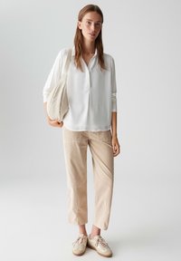 Weiße Bluse mit langen Ärmeln und Henley-Ausschnitt, kombiniert mit hellbeigen Hosen und braunen Sneakern. Das Modell hält eine cremefarbene Tragetasche.