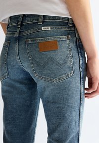 Person trägt blaue Denim Wrangler Jeans mit sichtbarer Gesäßtasche und braunem Leder Wrangler-Label, steht mit rechter Hand locker an der Seite.