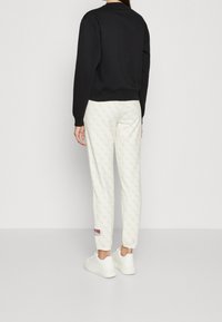 Sudadera negra con cuello redondo, combinada con pantalones de chándal crema que tienen un patrón sutil y un acento de logo rojo, completada con zapatillas blancas.