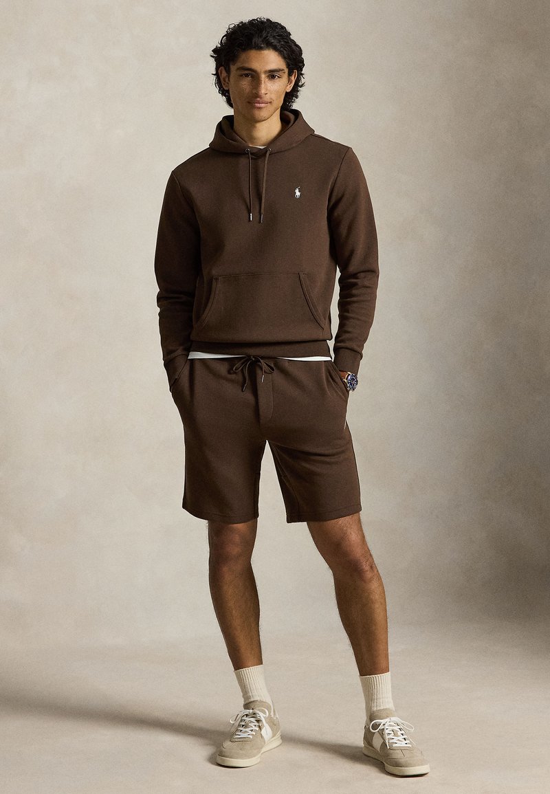 Ung mand står med hænderne i lommerne iført brun hættetrøje og matchende shorts, beige sneakers og hvide sokker mod en ensfarvet baggrund.