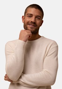 Hellbeige, strukturierter Strickpullover mit Rundhalsausschnitt und gerippten Bündchen. Hat eine lockere Passform und wird von einem Mann mit kurzem Haar getragen.