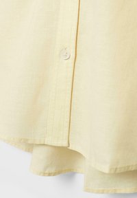 Tissu jaune avec une rangée verticale de boutons, montrant les coutures détaillées et l'ourlet superposé d'une chemise ou d'un chemisier.