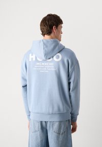Lichtblauwe hoodie met een groot wit logo en tekst op de achterkant, afgewerkt met een ribgebreide onderrand en manchetten.
