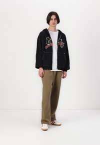 Svart hettegenser med røde og hvite logoer, kombinert med beige bukser og hvite joggesko med røde striper. Enkelt design, avslappet passform.